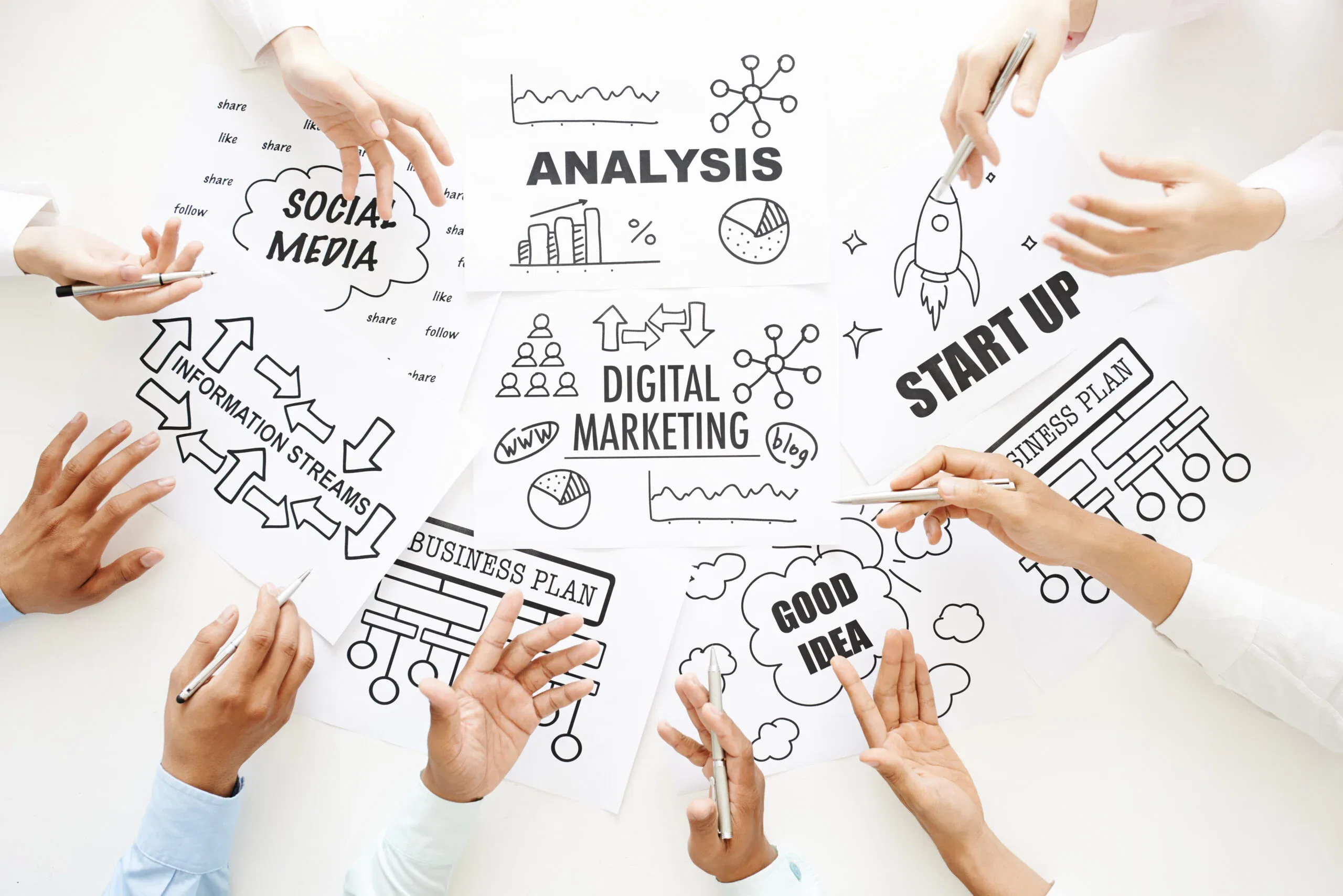 A top-down view of a marketing team’s hands pointing to sketches of business plans, social media icons, and digital marketing diagrams on a white table.