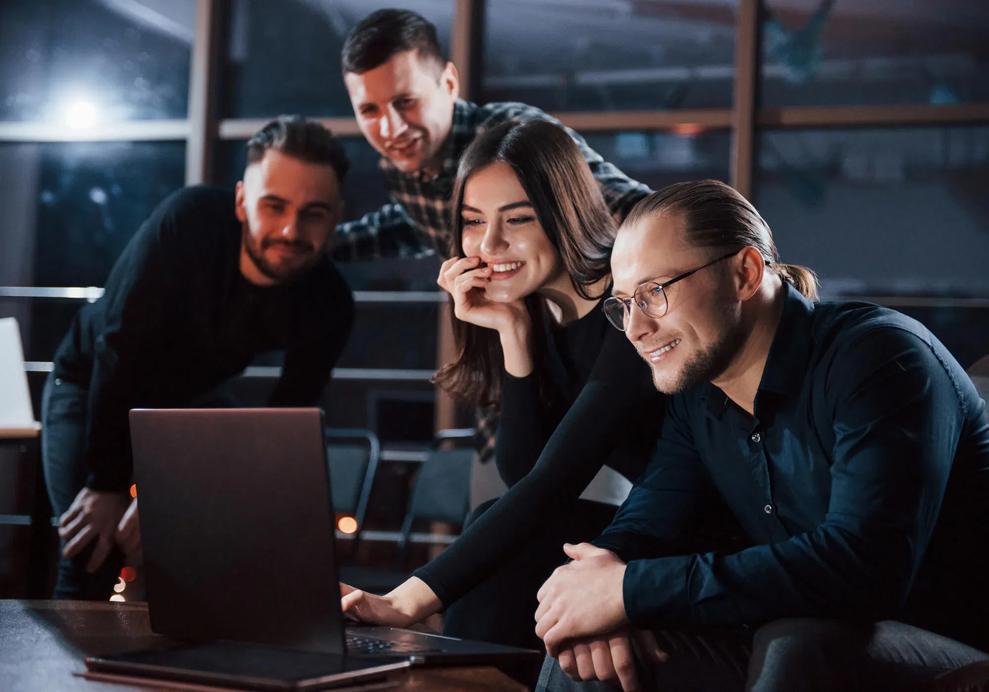 A group of four diverse young professionals smiling and looking at a laptop screen during a late-evening collaborative strategy session in a modern office.