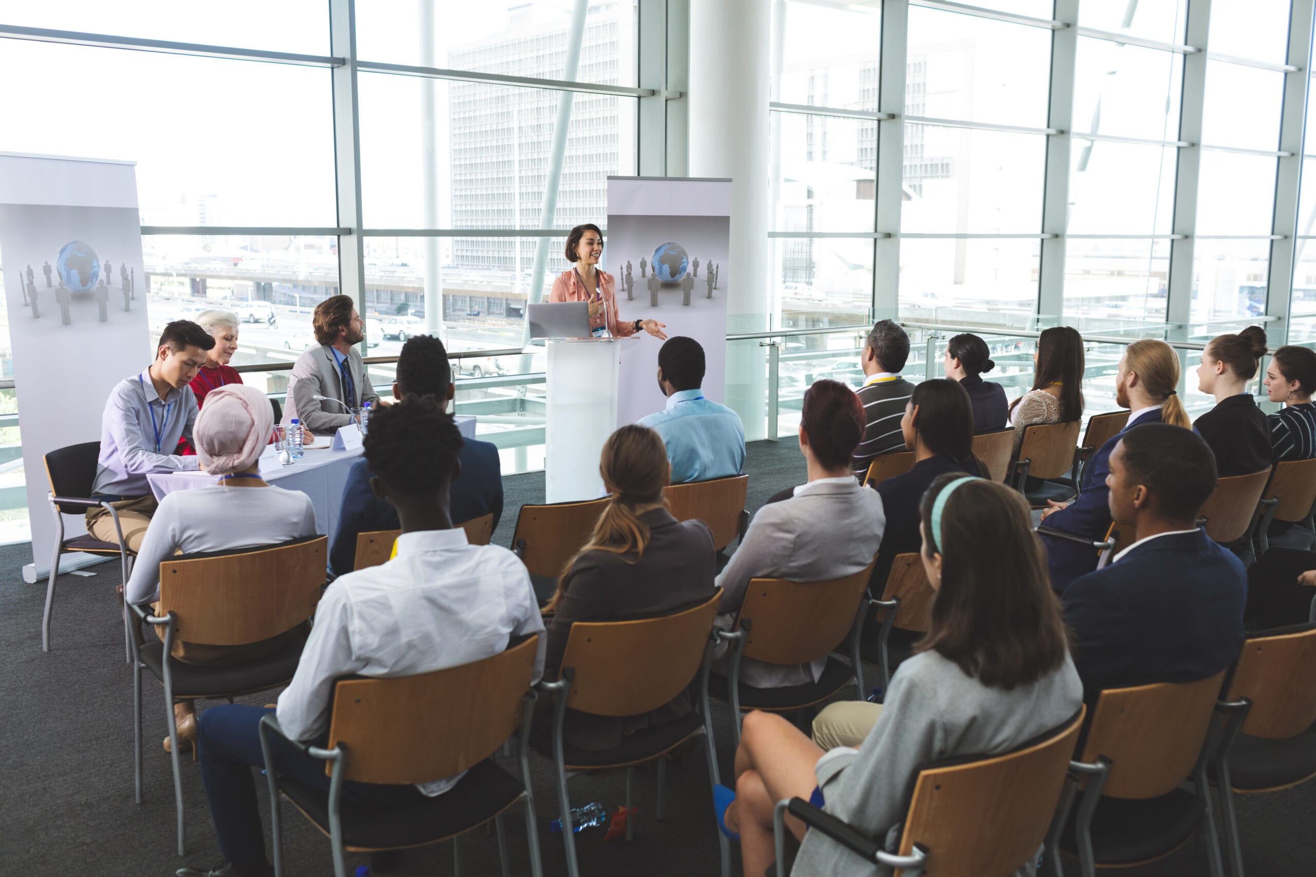 A professional businesswoman presenting to a diverse audience in a modern, sunlit conference hall with large floor-to-ceiling windows and presentation banners.