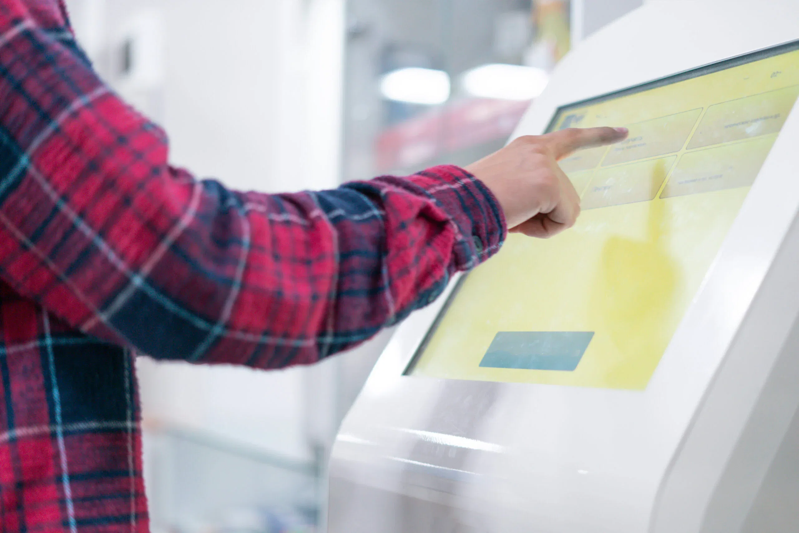 A side profile shot of a person in a red and blue plaid shirt using a finger to interact with a bright yellow touch-screen display on a white digital kiosk.
