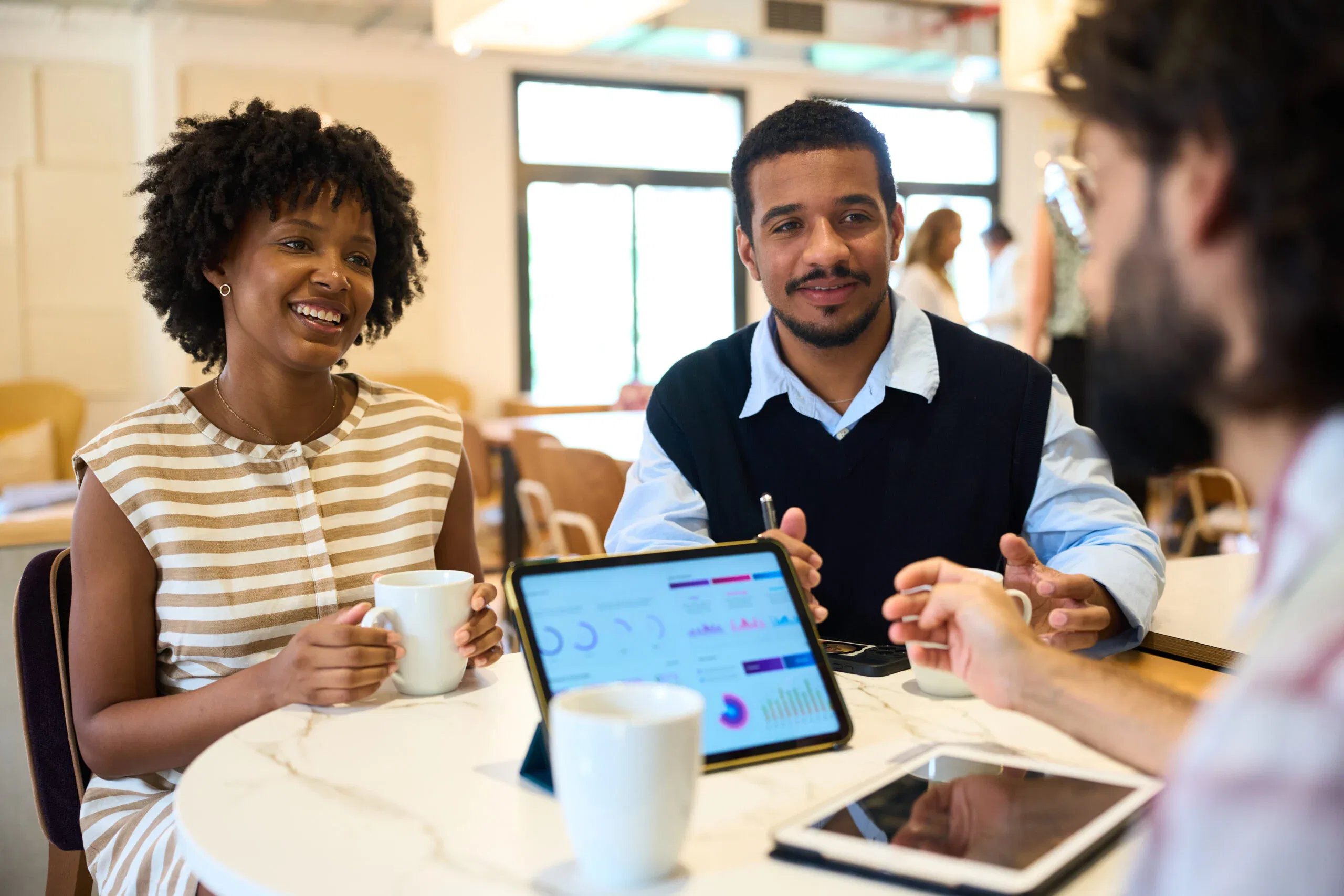 A diverse group of professionals sitting at a marble table in a bright office, discussing data visualizations on a tablet while holding coffee mugs.