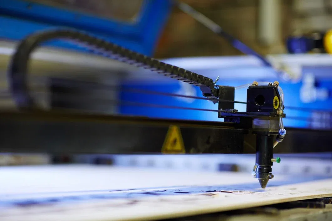 A close-up side view of a CNC laser cutting head in a blue-lit industrial workshop, carving intricate patterns into a white substrate.