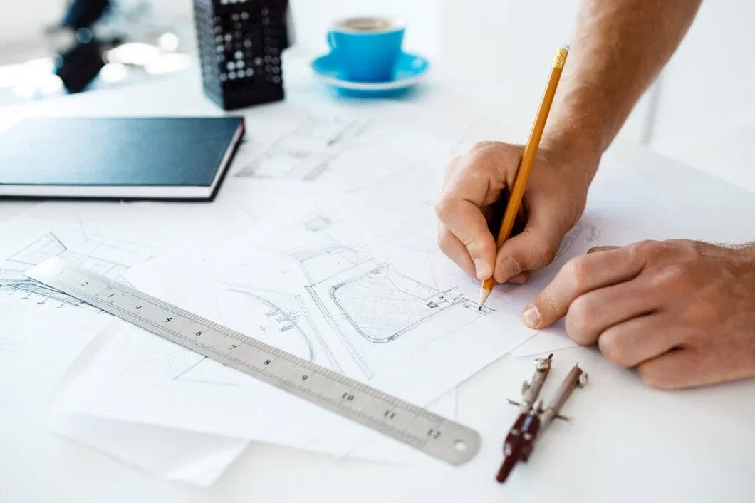 A close-up of a designer's hands using a pencil, ruler, and compass to draw detailed technical sketches on white paper in a modern office.