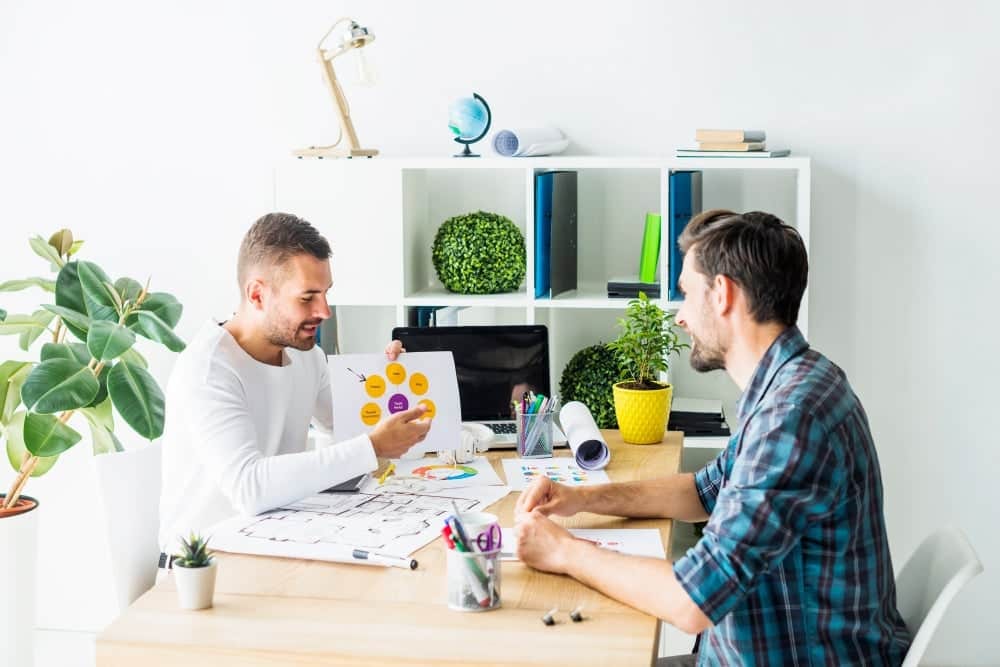 Two men in a bright office discussing architectural floor plans and a circular 