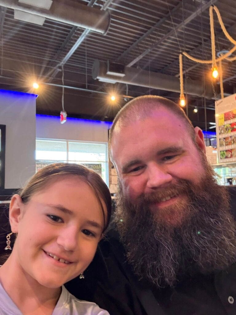 A smiling man with a full brown beard and a young girl posing for a close-up selfie inside a restaurant with industrial ceilings and hanging Edison bulbs.