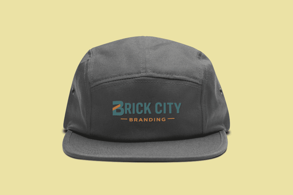 A dark charcoal 5-panel camper-style hat featuring the Brick City Branding logo embroidered on the front panel, set against a pale yellow background.