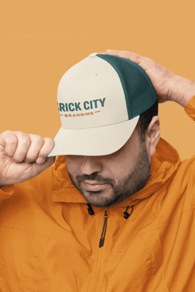 A man with a beard and an orange jacket adjusting a cream and teal mesh trucker hat featuring the Brick City Branding logo.