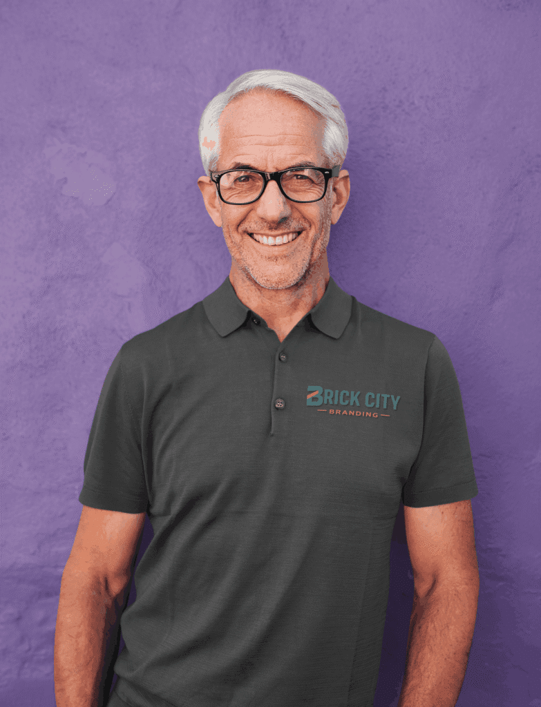 A smiling older man with grey hair and glasses wearing a charcoal grey polo shirt with the Brick City Branding logo on the left chest, posing against a purple wall.