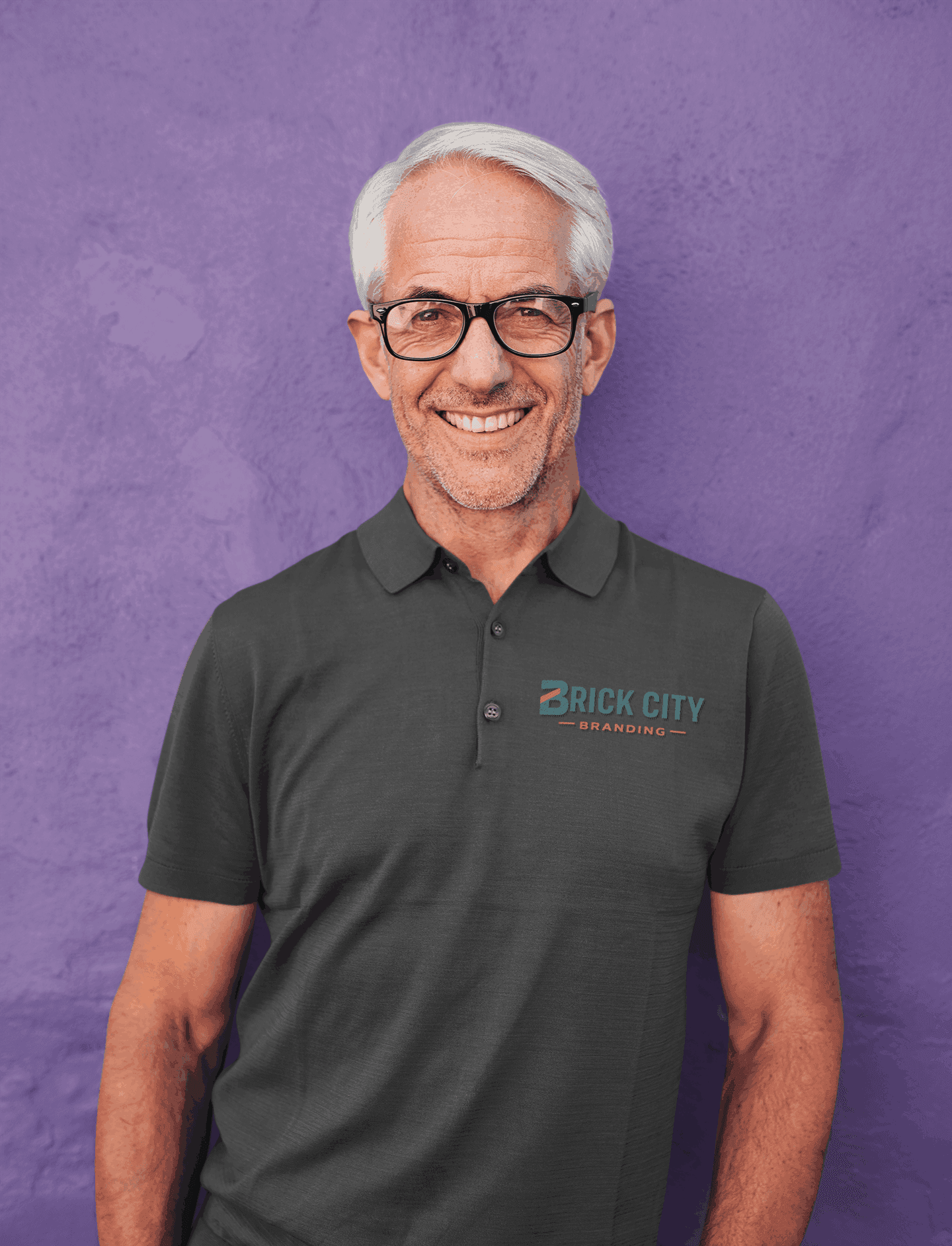 A smiling older man with grey hair and glasses wearing a charcoal grey polo shirt with the Brick City Branding logo on the left chest, posing against a purple wall.