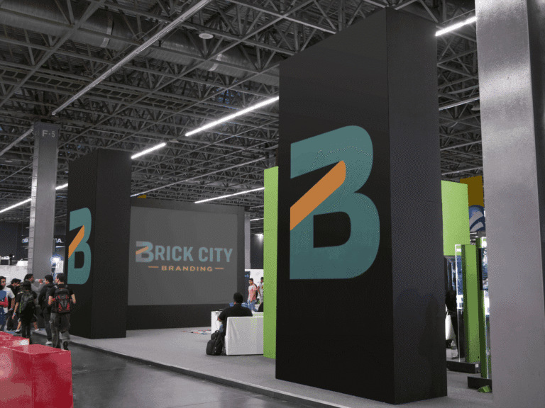 A large modern trade show exhibit for Brick City Branding featuring tall black monolith pillars with teal and orange logos and a large digital screen displaying the full brand name.
