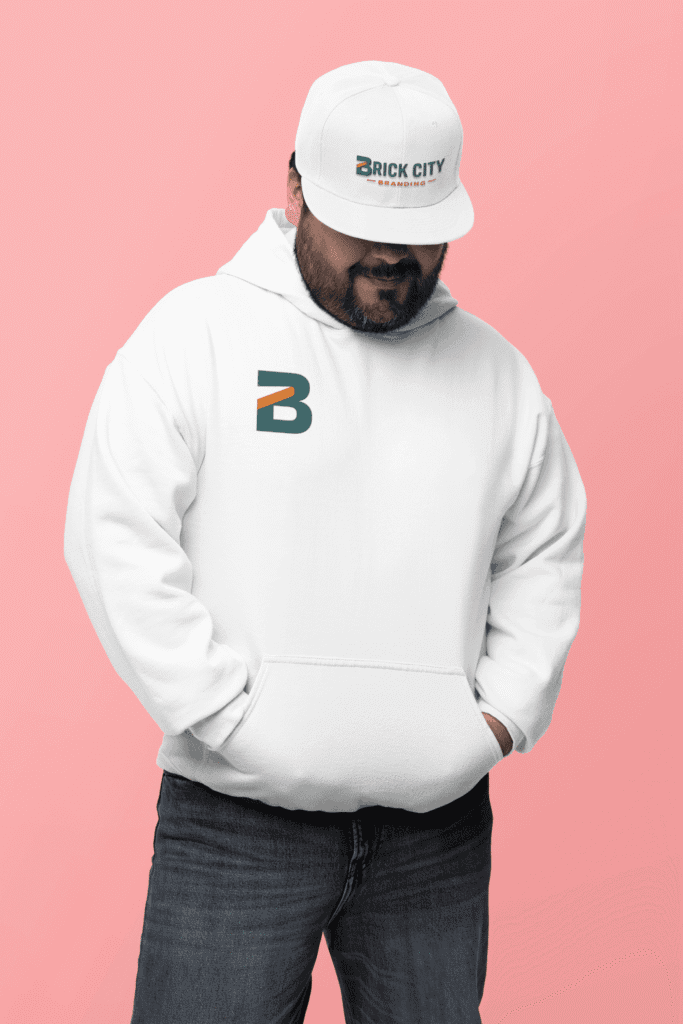 A man with a beard wearing a white Brick City Branding hoodie and a matching white flat-brim cap against a soft pink background.