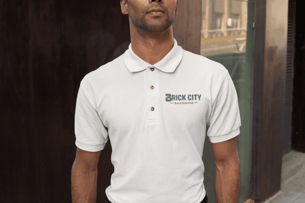 A man wearing a white custom polo shirt with the Brick City Branding logo embroidered on the left chest, standing in an urban outdoor setting.