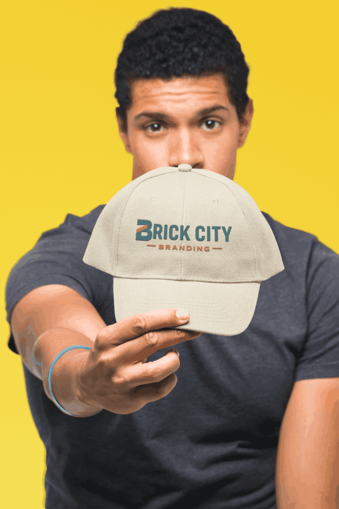 A white 6-panel structured baseball cap featuring the Brick City Branding logo embroidered on the front, displayed at a side-profile angle against a solid orange background.