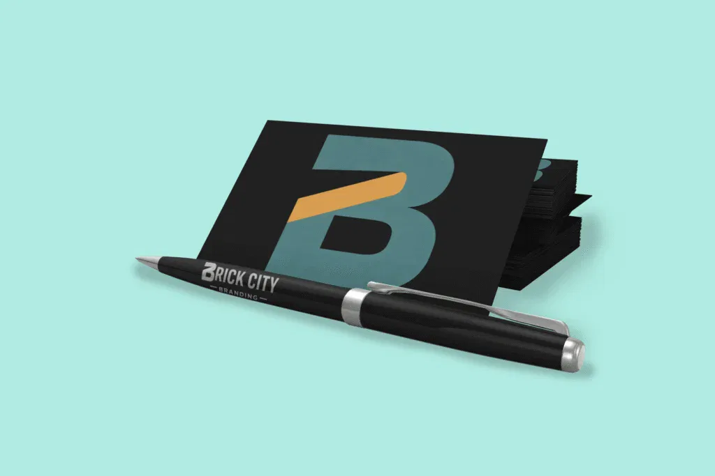 A premium black metal ballpoint pen with silver accents lying in front of a stack of matte black business cards featuring the Brick City Branding logo.