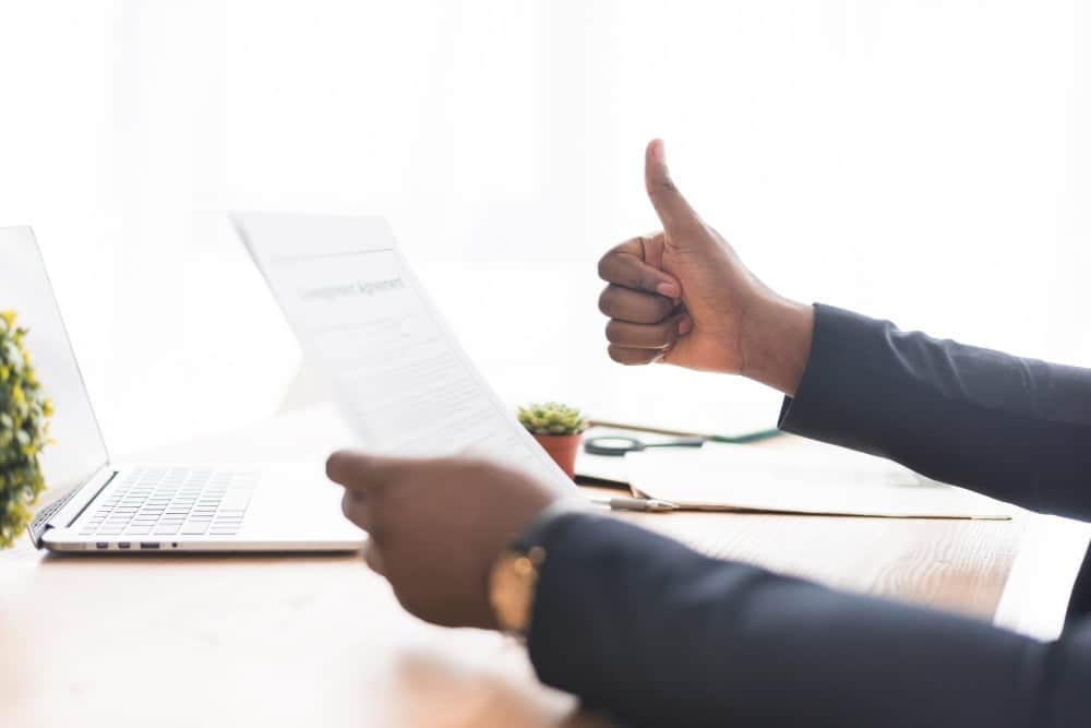 Close-up of a professional in a suit holding a signed agreement document and giving a thumbs up at a bright office desk with a laptop.