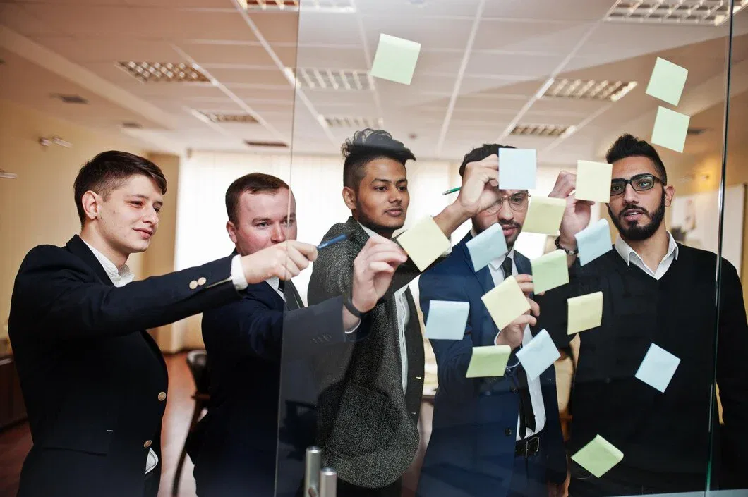 A group of five professional marketing strategists in business attire placing post-it notes on a glass wall during a brand development workshop.