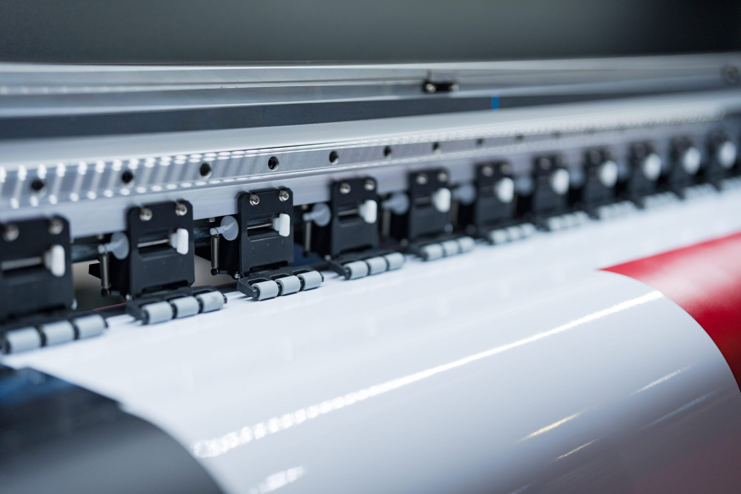 A close-up of a high-speed industrial wide-format printer feeding a roll of white vinyl with red graphic elements through precision rollers.