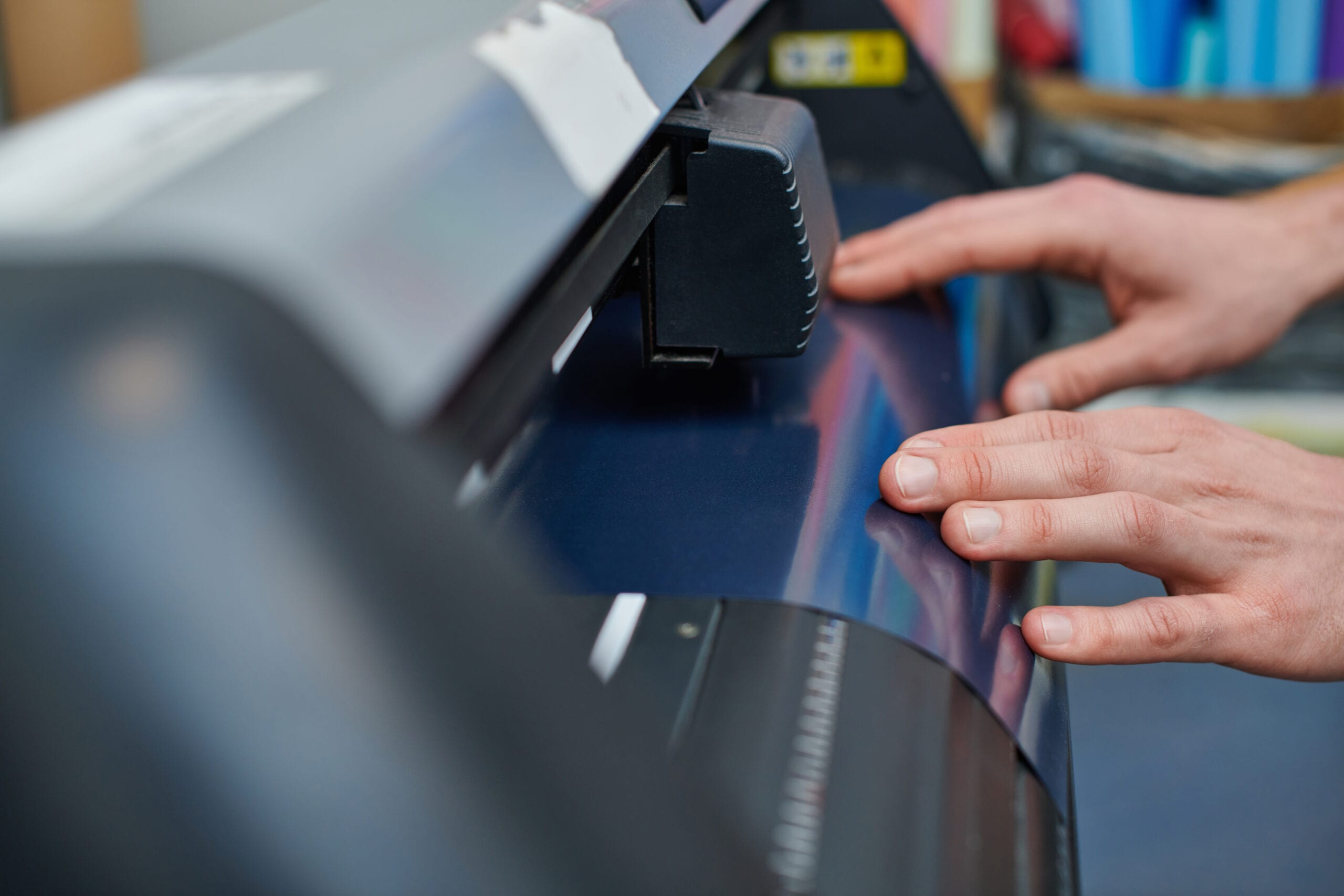 A close-up of a person's hands guiding blue vinyl through a professional cutting plotter machine in a production studio.