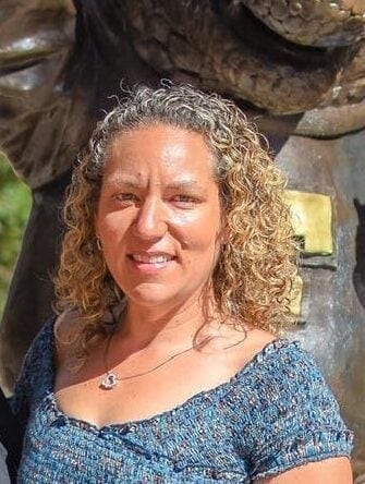 A smiling woman with shoulder-length curly blonde hair wearing a blue smocked top and a heart necklace, posing outdoors.