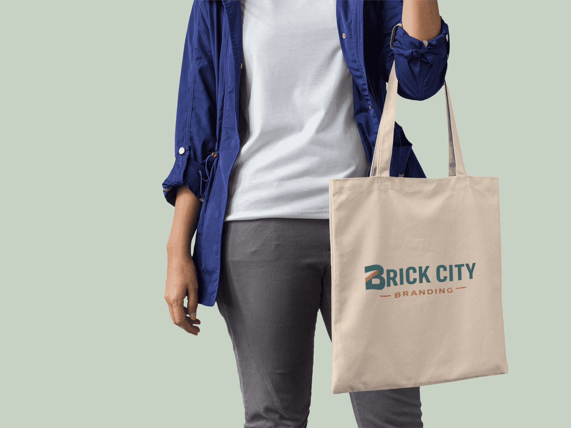A person holding a natural cotton canvas tote bag featuring the Brick City Branding logo in teal and orange.