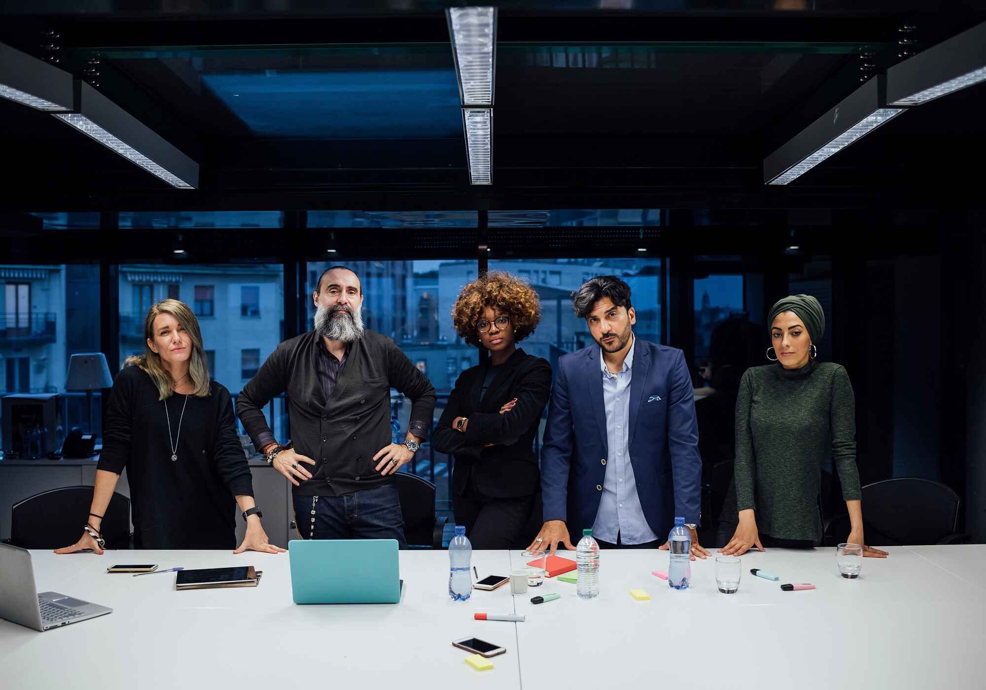A diverse group of five confident business partners standing behind a conference table in a modern office at dusk.