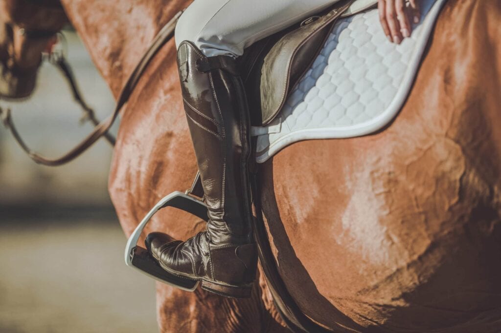 Close-up of a rider's tall brown leather boot in a silver stirrup, showing the detail of the saddle and white quilted saddle pad on a chestnut horse.