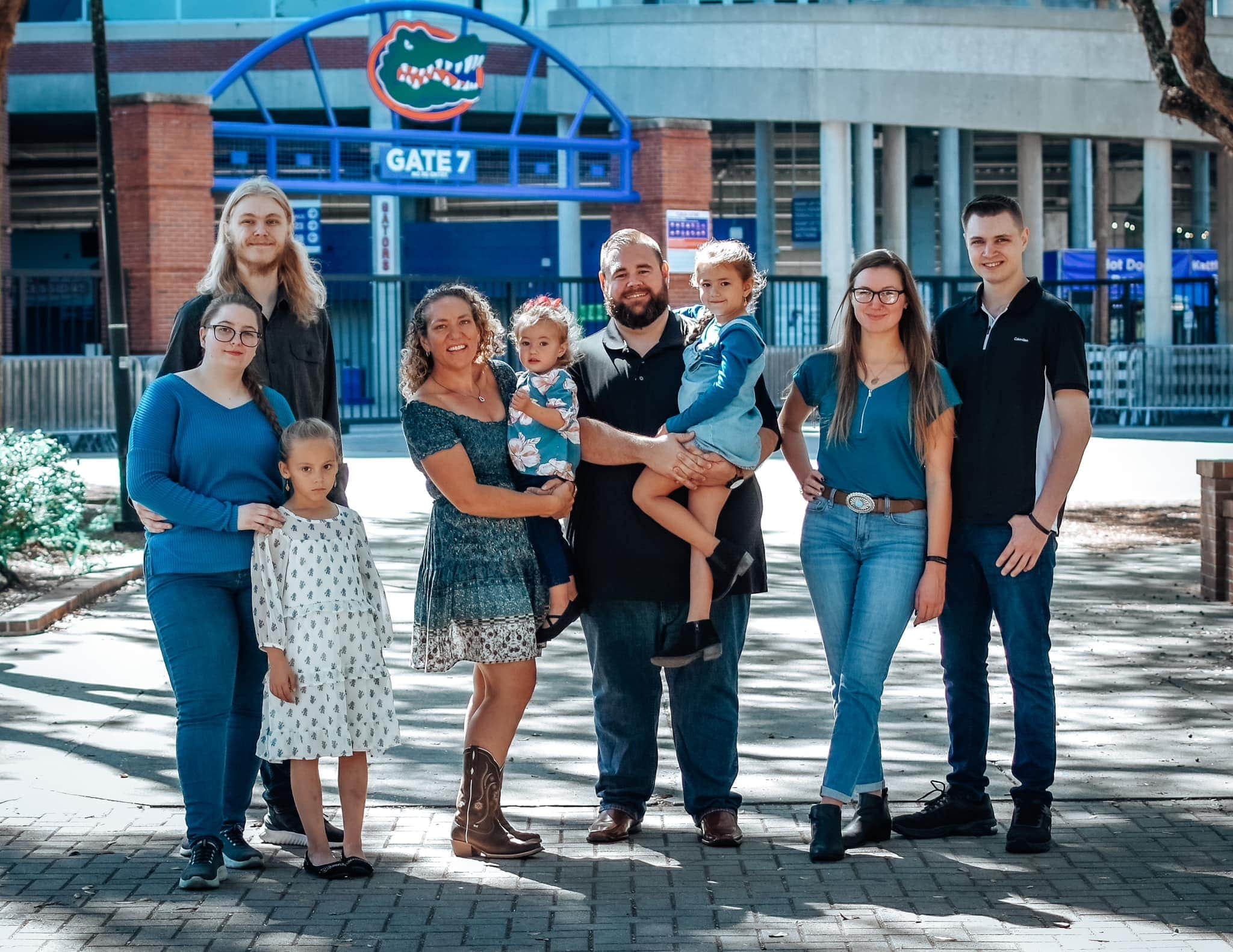 A large family of eight, including children and adults, posing together in front of Gate 7 at the Ben Hill Griffin Stadium, featuring the Florida Gators logo.