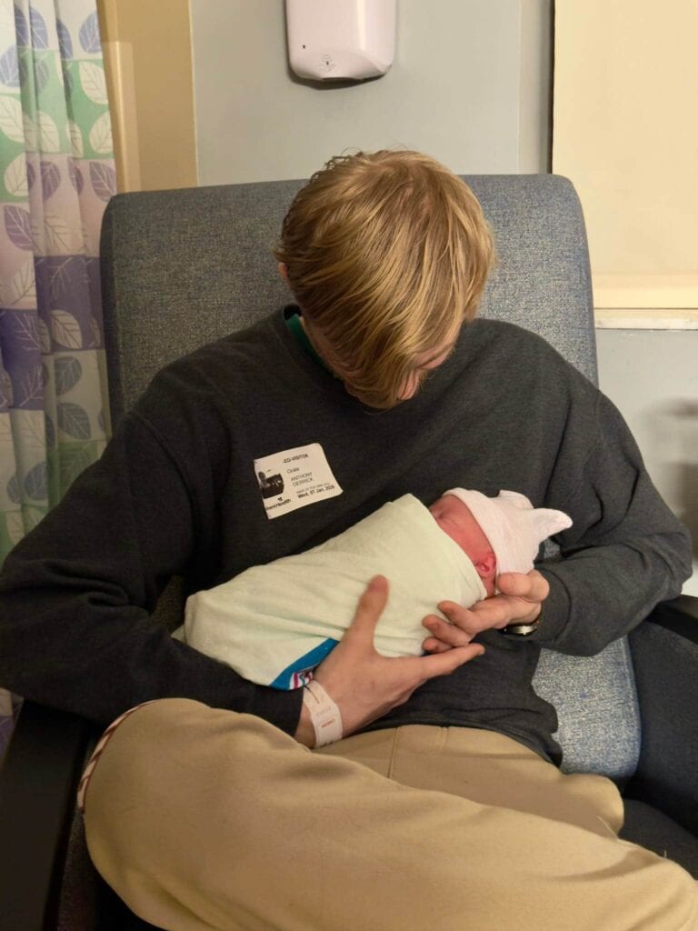 A young man with blonde hair sitting in a hospital chair, looking down lovingly while holding a swaddled newborn baby wearing a white knit cap.