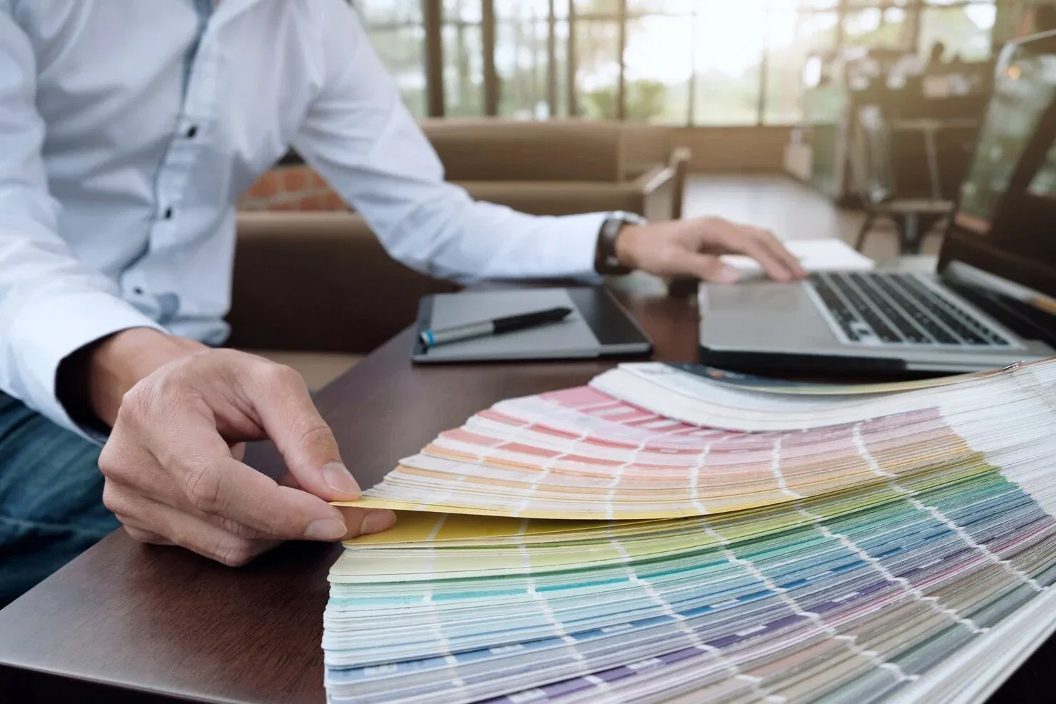 Close-up of a designer's hands browsing through a large fan of color swatches on a wooden desk next to a laptop and digital drawing tablet.