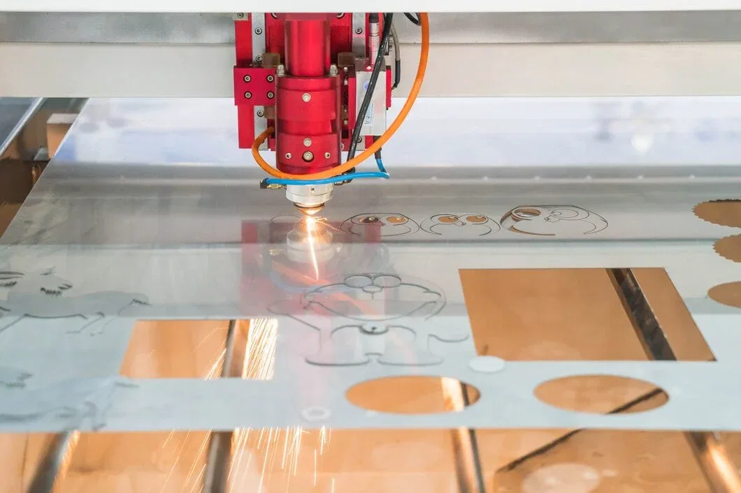 A red CNC laser cutting machine head firing a high-intensity beam through a metal sheet, creating bright sparks while cutting intricate shapes.