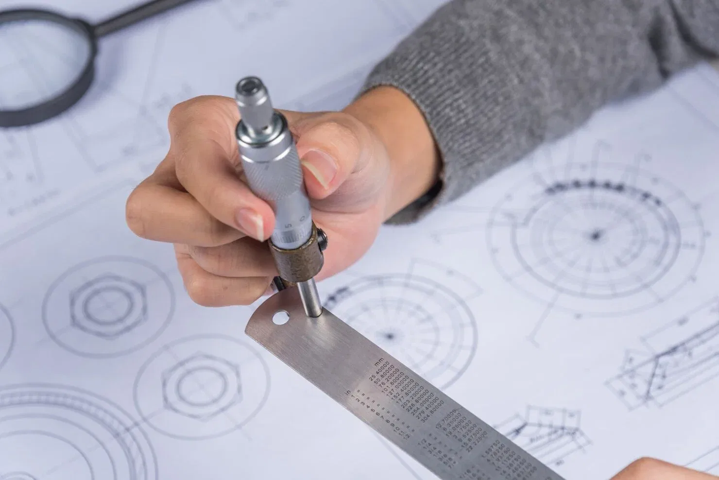 A close-up of hands using a micrometer and steel ruler to take precise measurements over detailed technical architectural blueprints.
