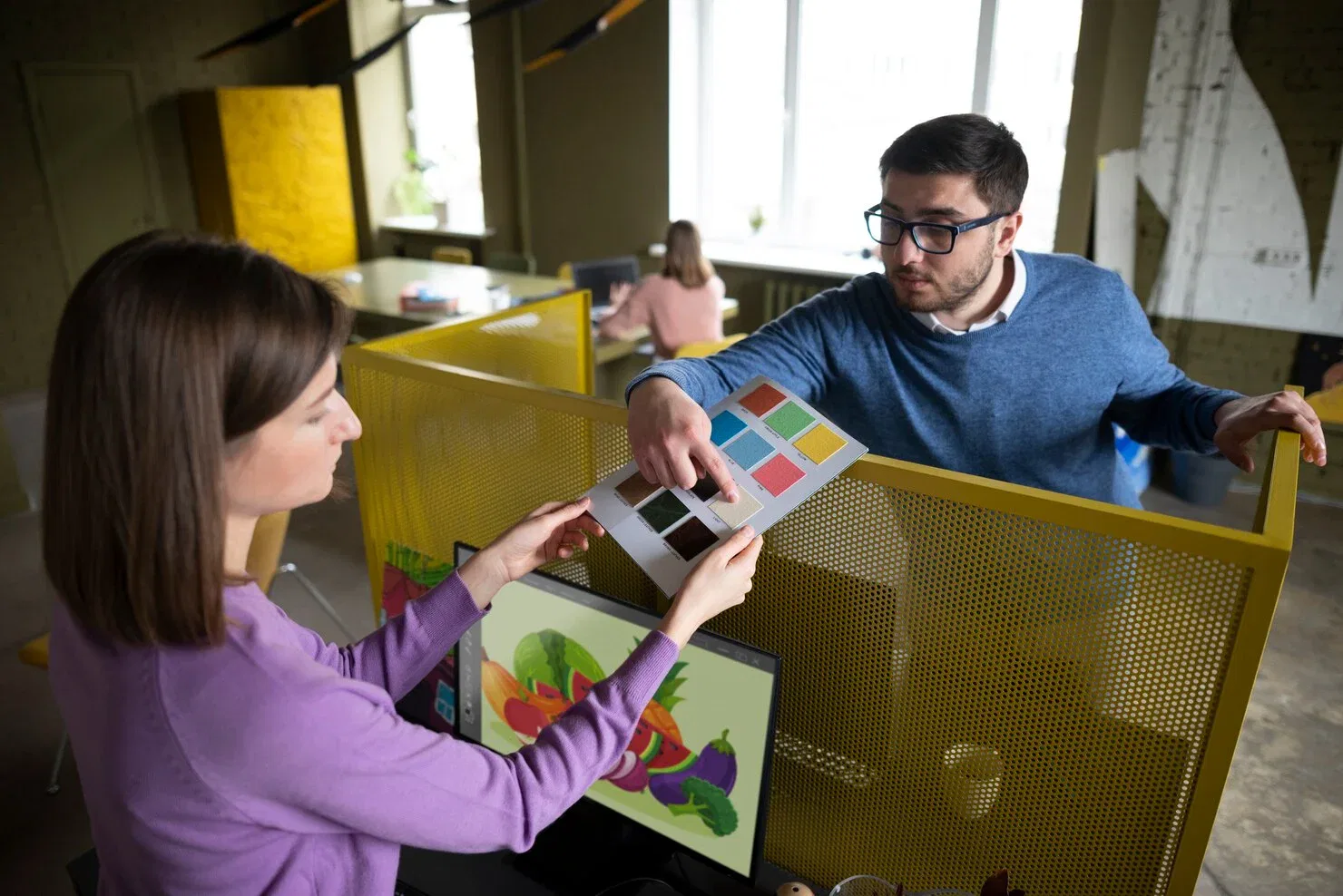 Two colleagues in a modern office with yellow partitions discussing a color palette while looking at a monitor displaying a vibrant vegetable illustration.