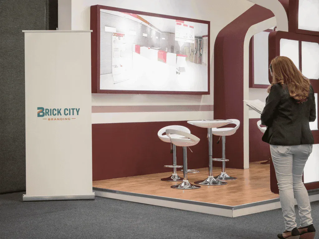 A vertical white retractable pull-up banner with the Brick City Branding logo positioned in a modern trade show booth with maroon accents and a white cafe table.