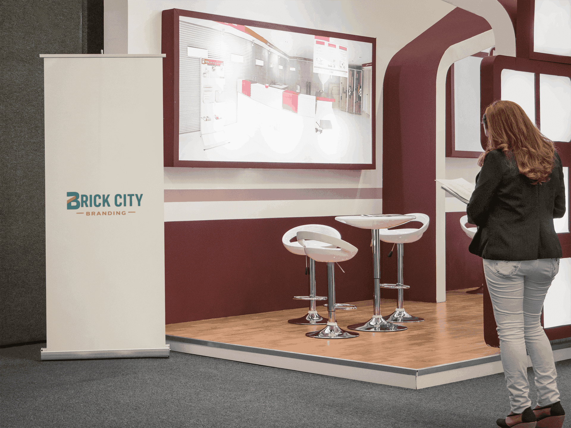 A vertical white retractable pull-up banner with the Brick City Branding logo positioned in a modern trade show booth with maroon accents and a white cafe table.