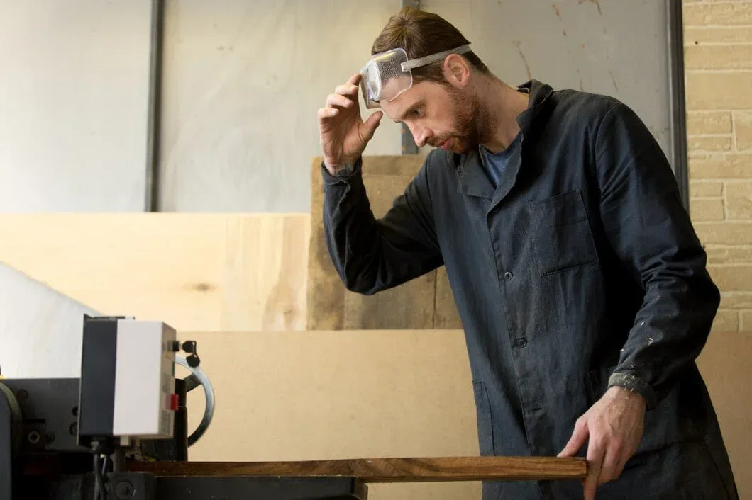 A professional carpenter wearing safety goggles meticulously operating a sawmill machine to cut a large slab of natural wood in a workshop.