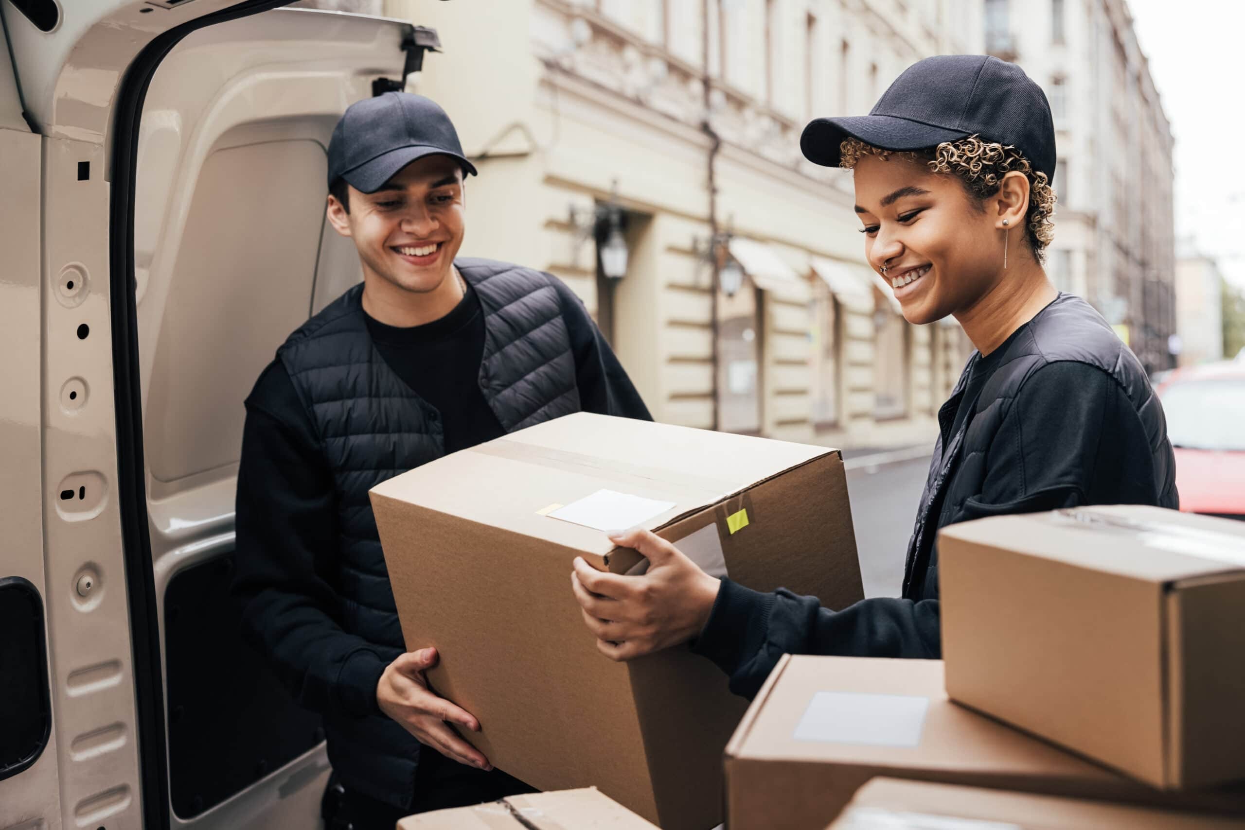 Two smiling couriers in black uniforms and caps unloading cardboard shipping boxes from the back of a white delivery van on a city street.