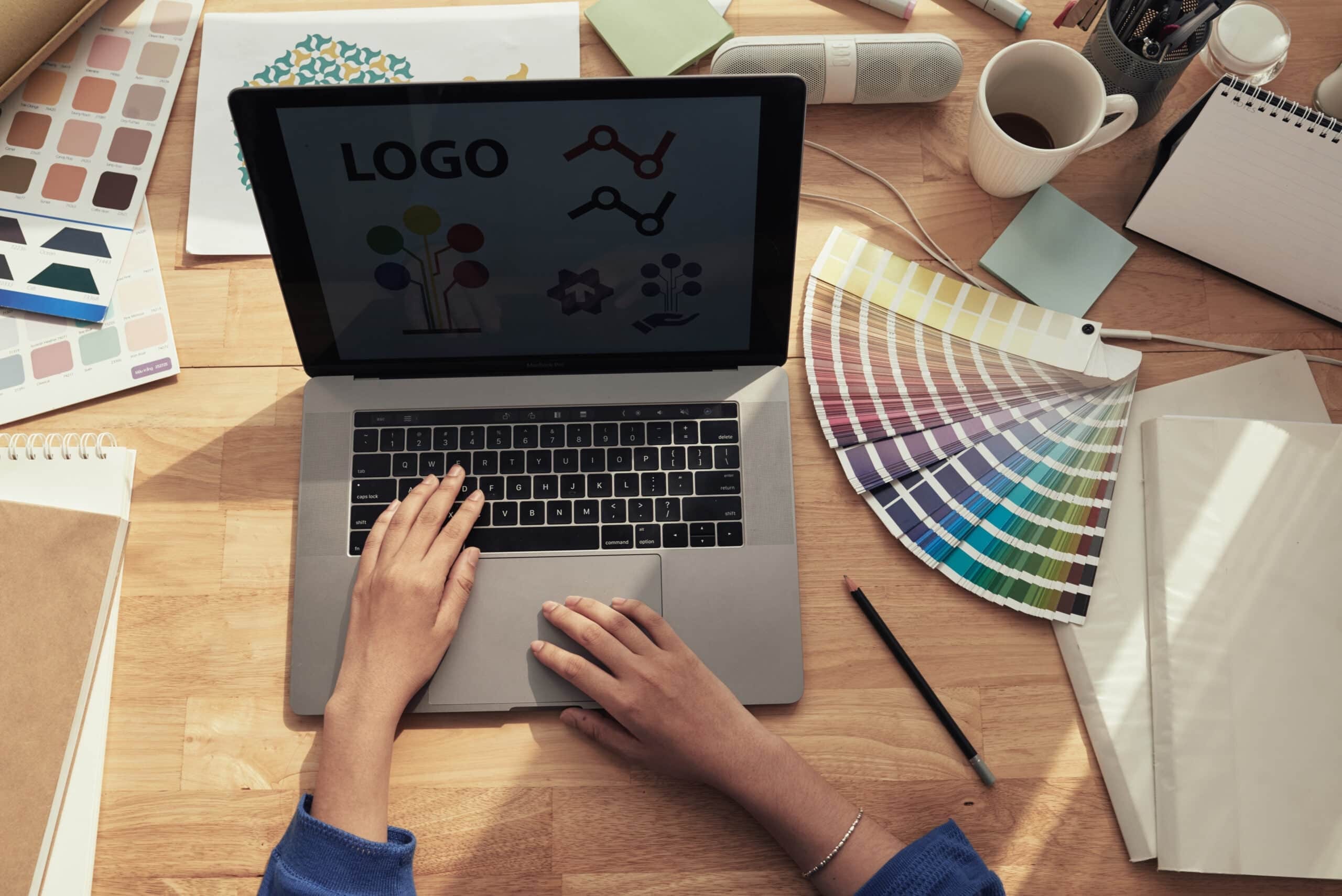 A top-down view of a designer working on a laptop with logo concepts on the screen, surrounded by Pantone color swatches and physical notebooks on a wooden desk.