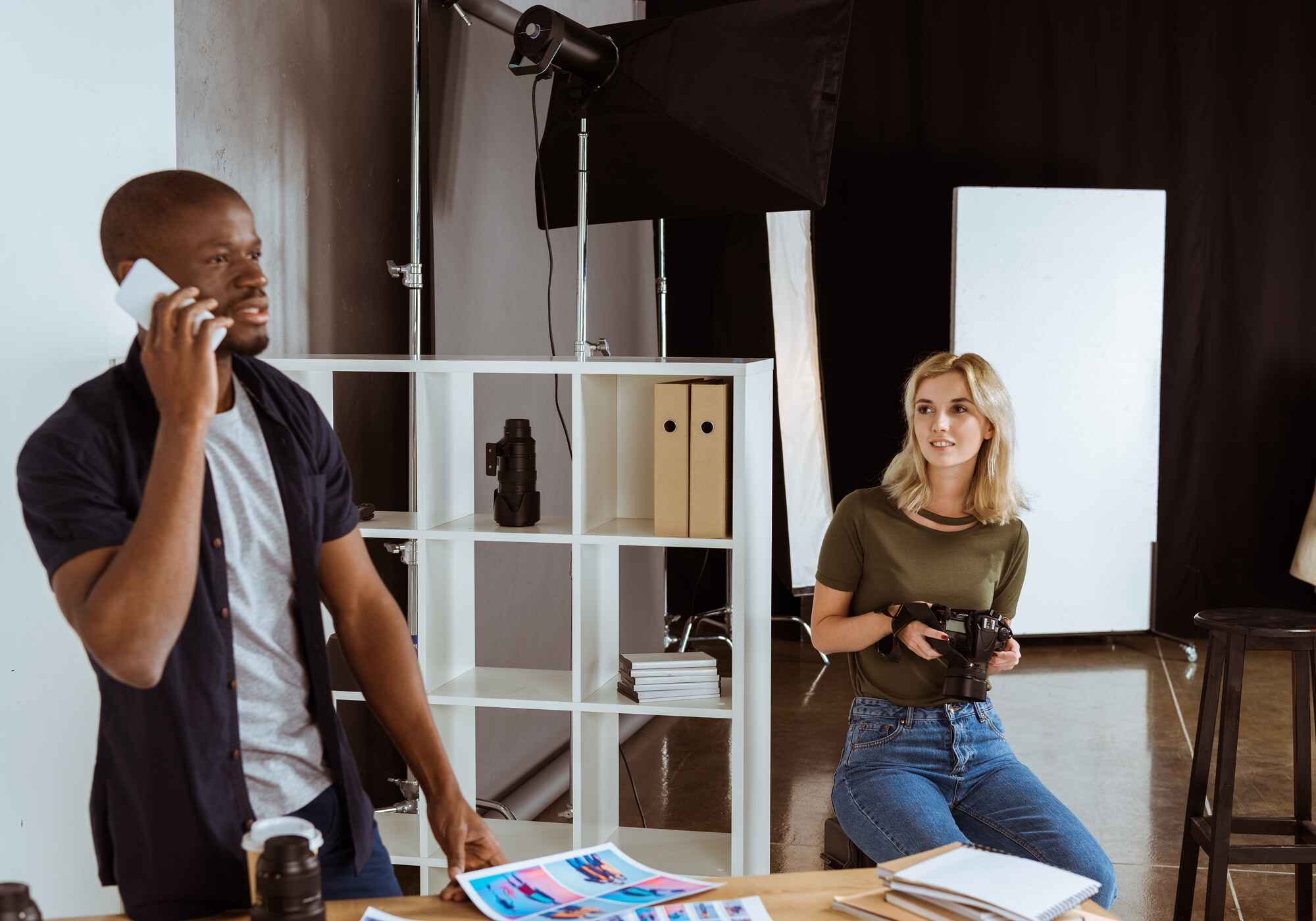 A male producer talking on a smartphone while a female photographer holds a camera in a professional studio with lighting equipment.
