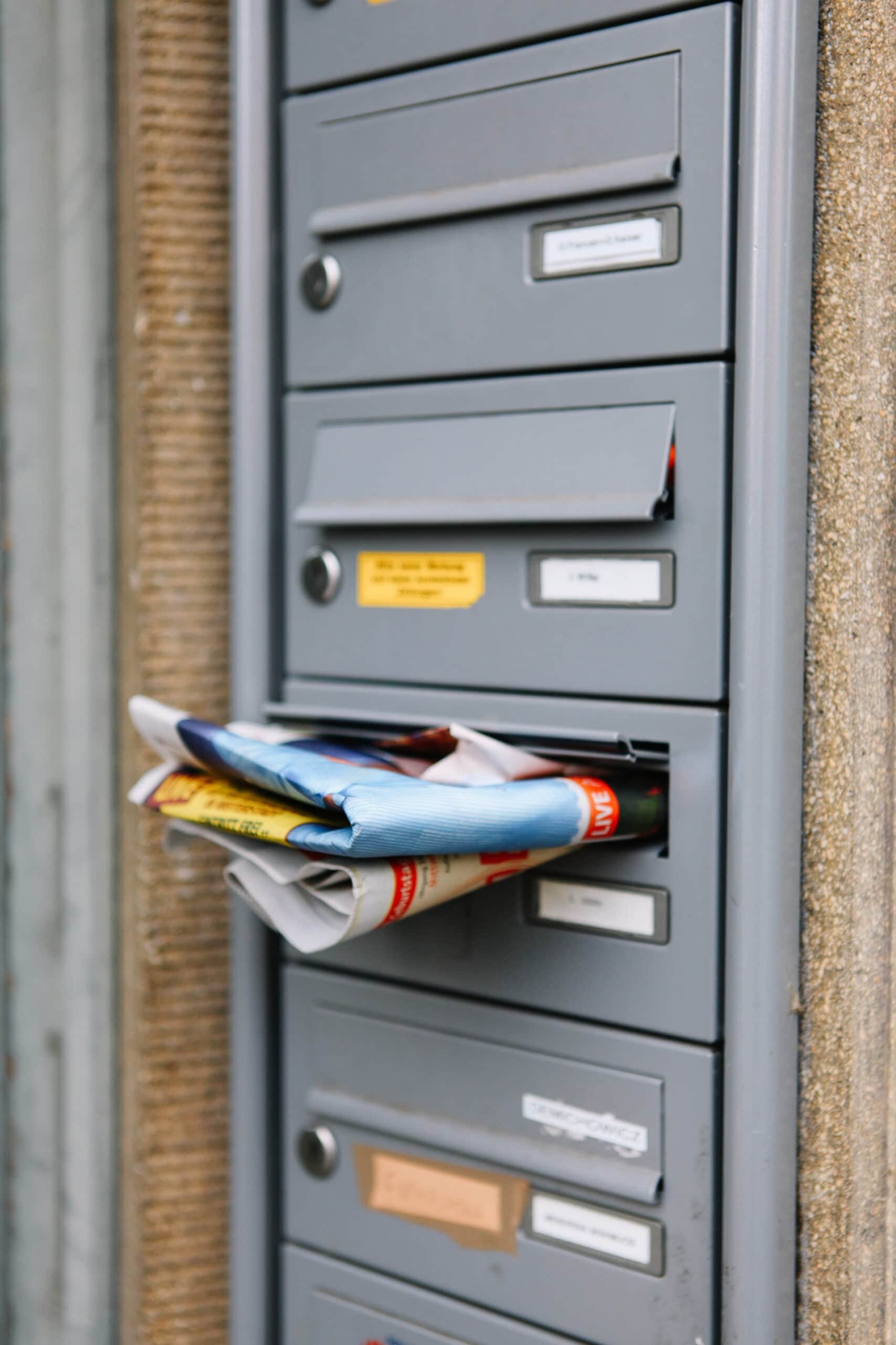 A vertical close-up of several gray apartment mailboxes, with one mailbox overflowing with folded newspapers and colorful direct mail flyers.