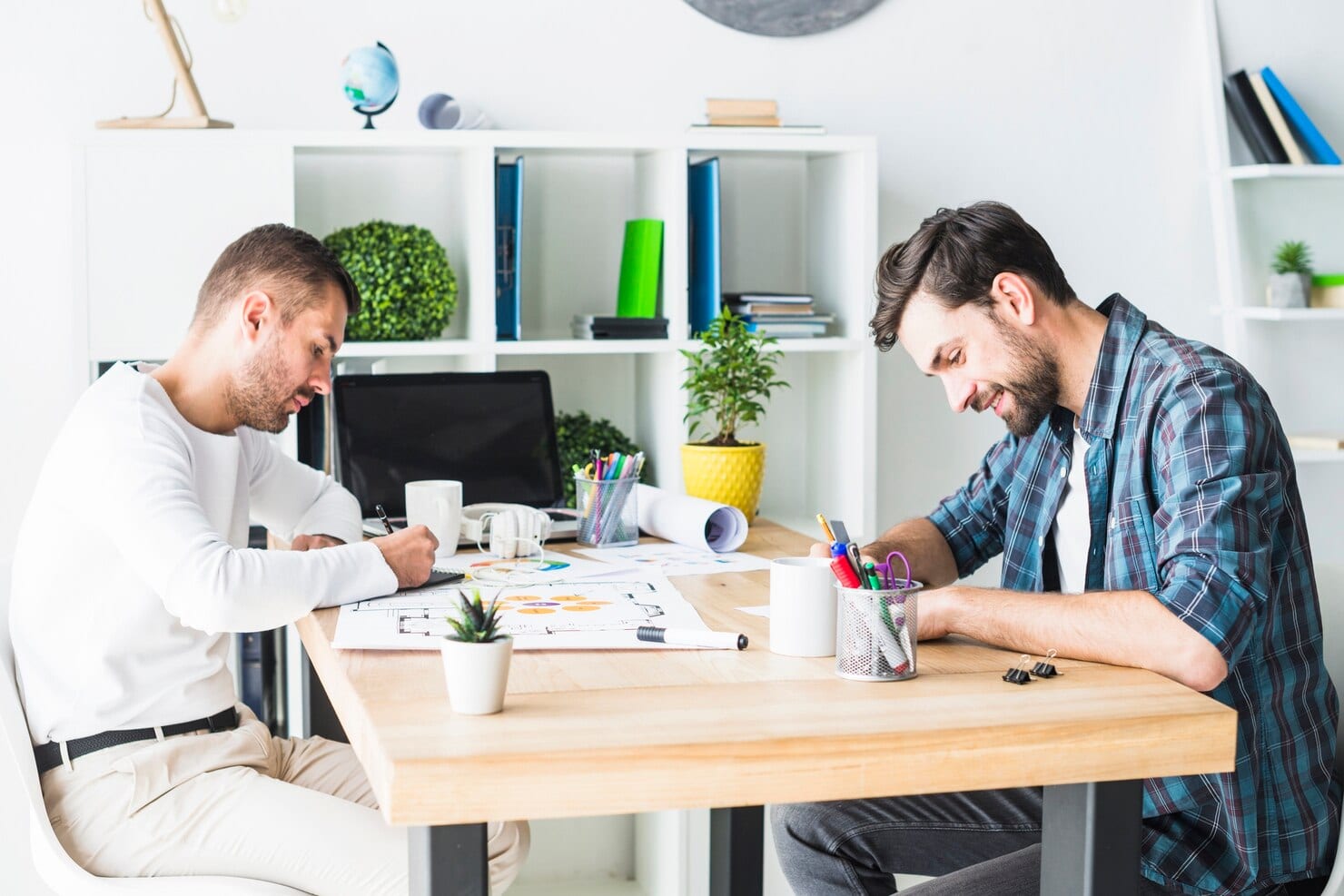 Two male graphic designers working diligently at a large wooden desk in a bright, modern office studio with creative tools and digital displays.