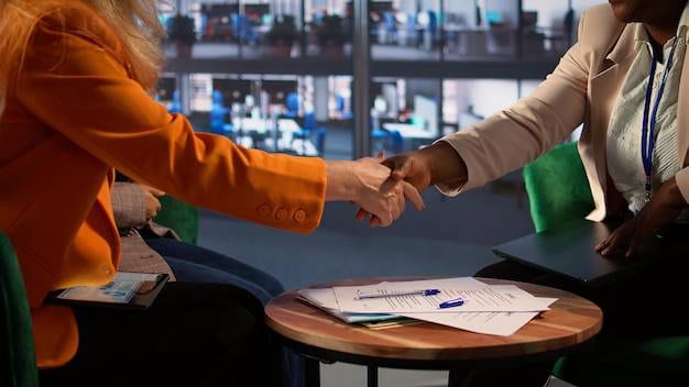Two professional women shaking hands over a wooden table with signed documents and pens in a modern office setting.