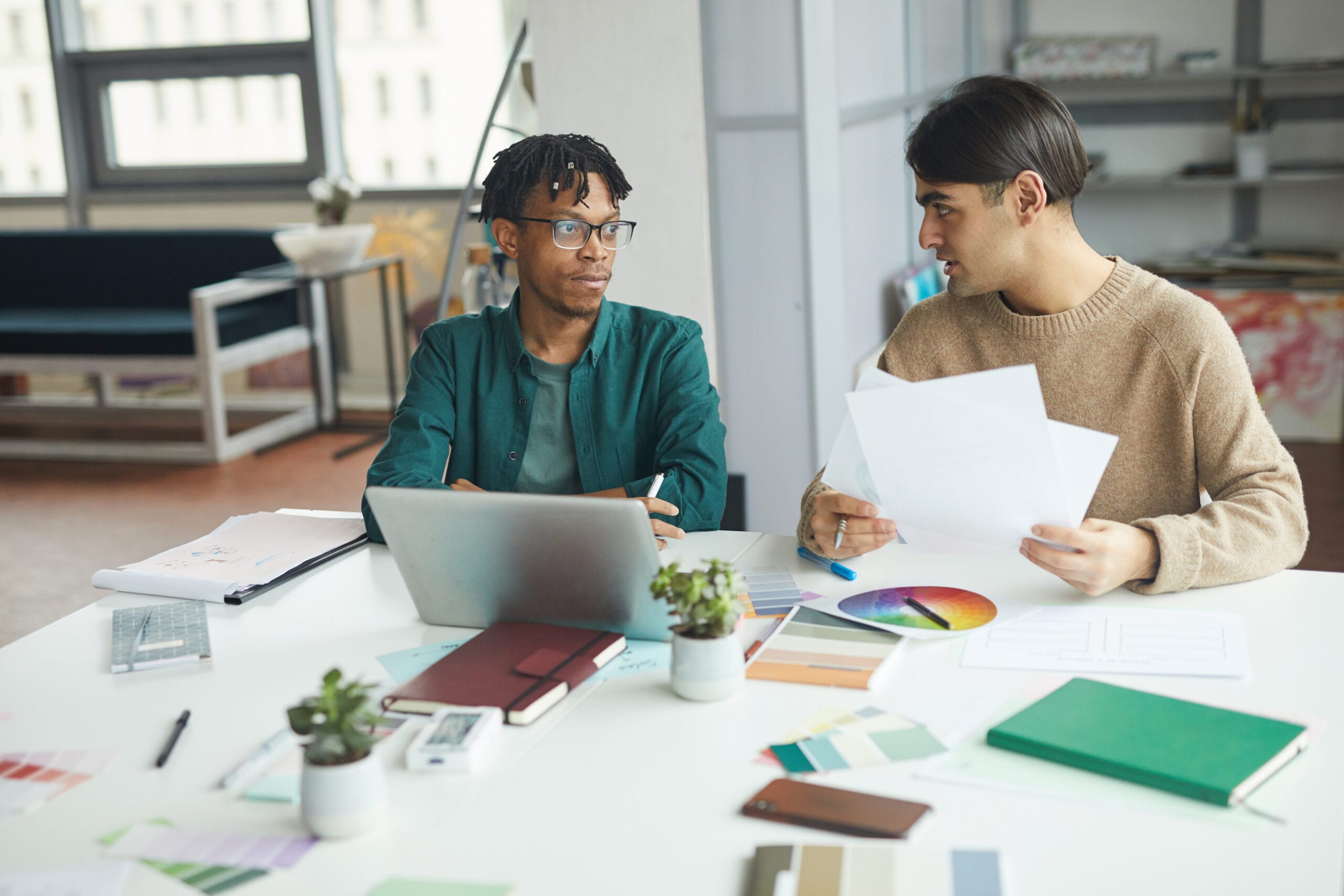 Two creative professionals in a bright studio discussing design documents at a desk with a laptop, color wheel, and swatch samples.