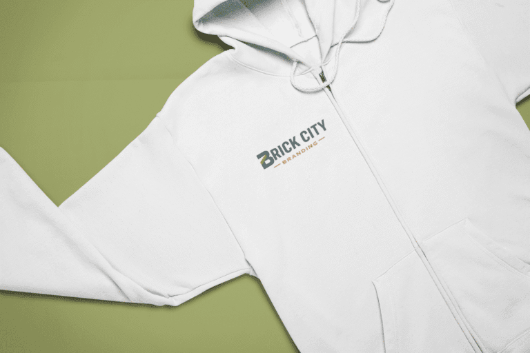 A top-down flat-lay view of a white full-zip hooded sweatshirt featuring the Brick City Branding logo on the left chest, placed on a split olive green and light green background.