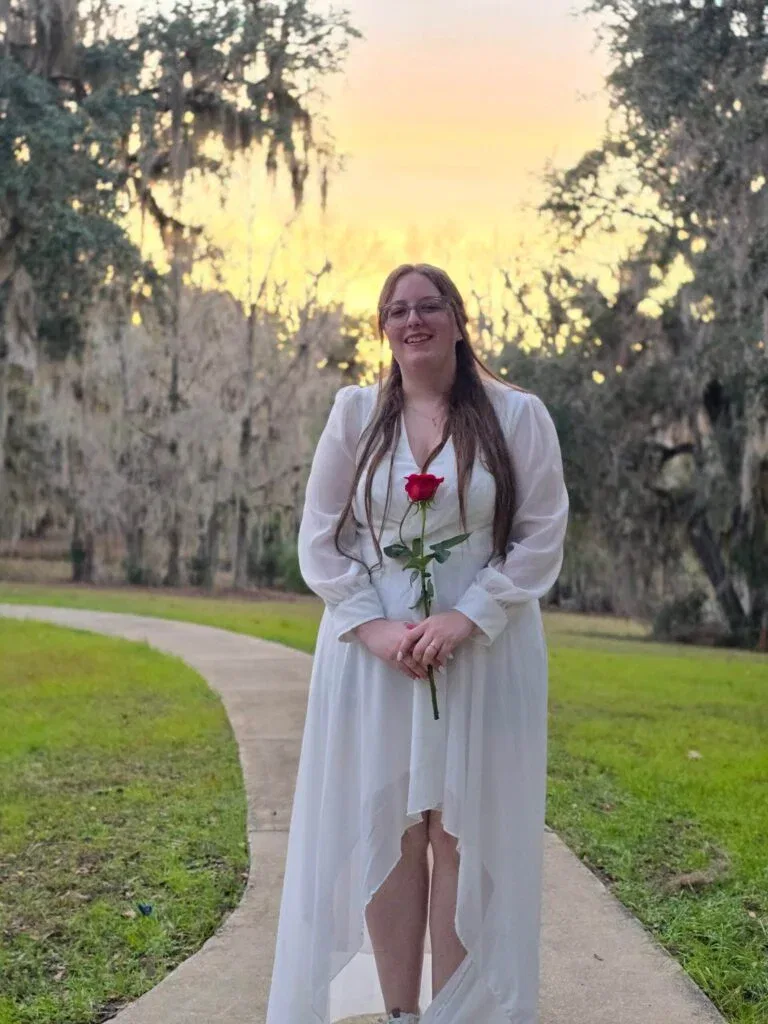 A smiling young woman in a flowy white high-low dress holding a single red rose, standing on a winding path under Spanish moss trees at sunset.