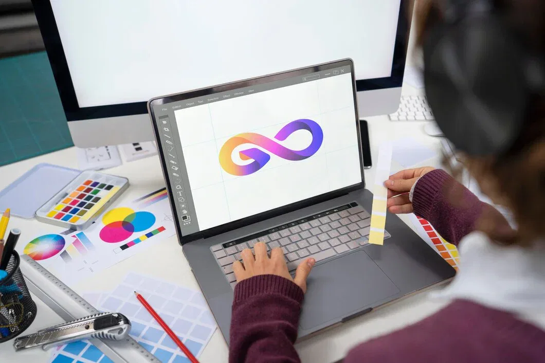 A graphic designer working on a laptop, creating a colorful 3D infinity loop logo, with color swatches and design tools visible on the desk.
