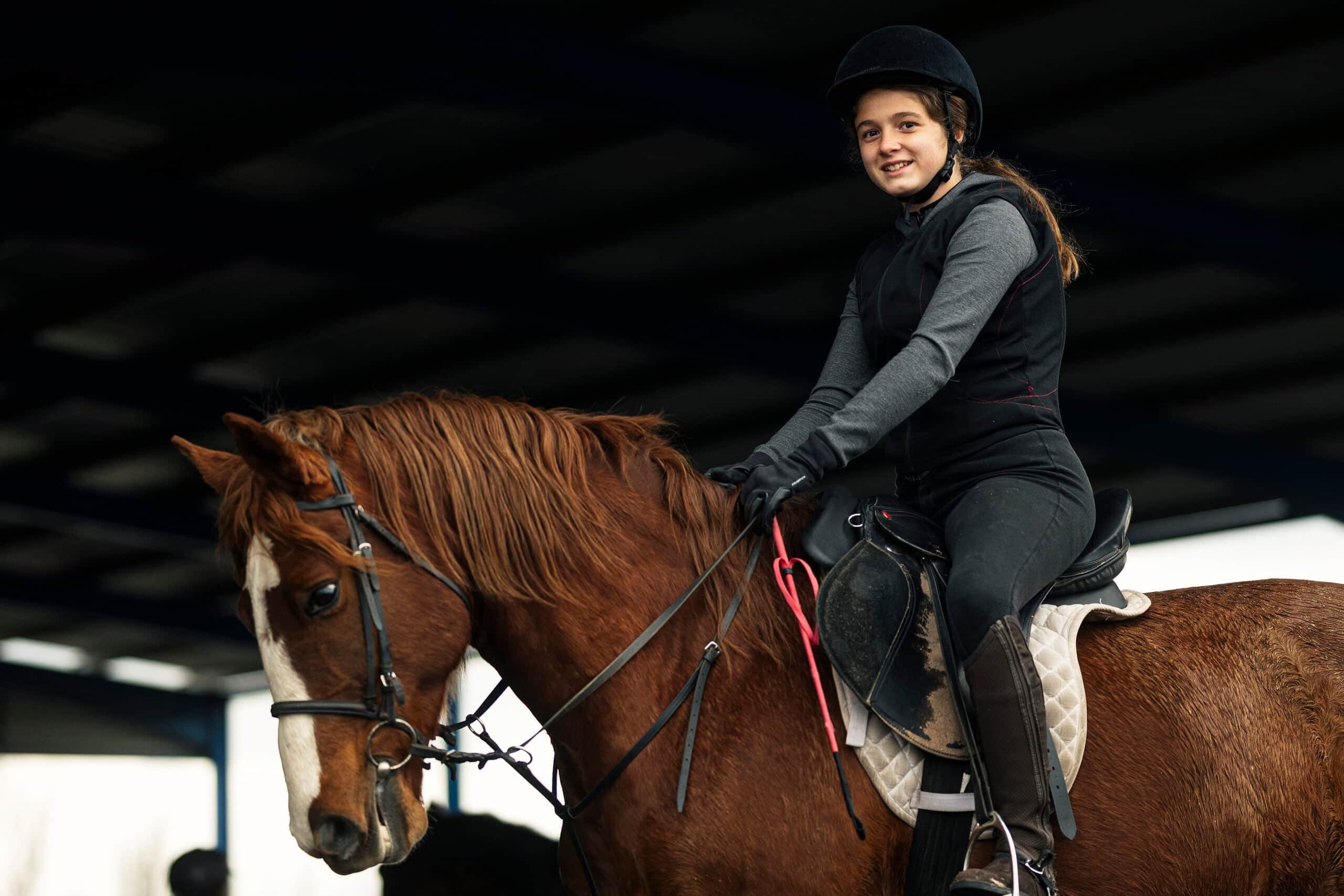 A young girl with a focused expression riding a brown horse, wearing a black equestrian helmet and a blue vest in an outdoor paddock.