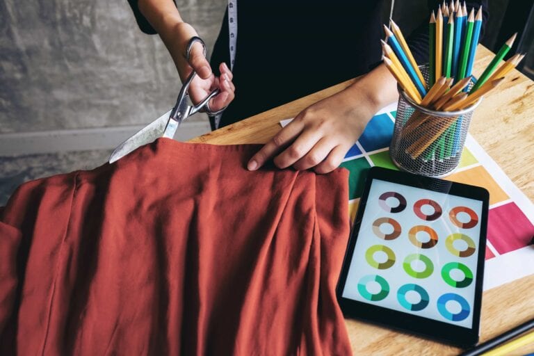 Top-down view of a dressmaker using professional shears to cut terracotta-colored fabric on a wooden desk, next to a tablet displaying color wheels and a cup of colored pencils.